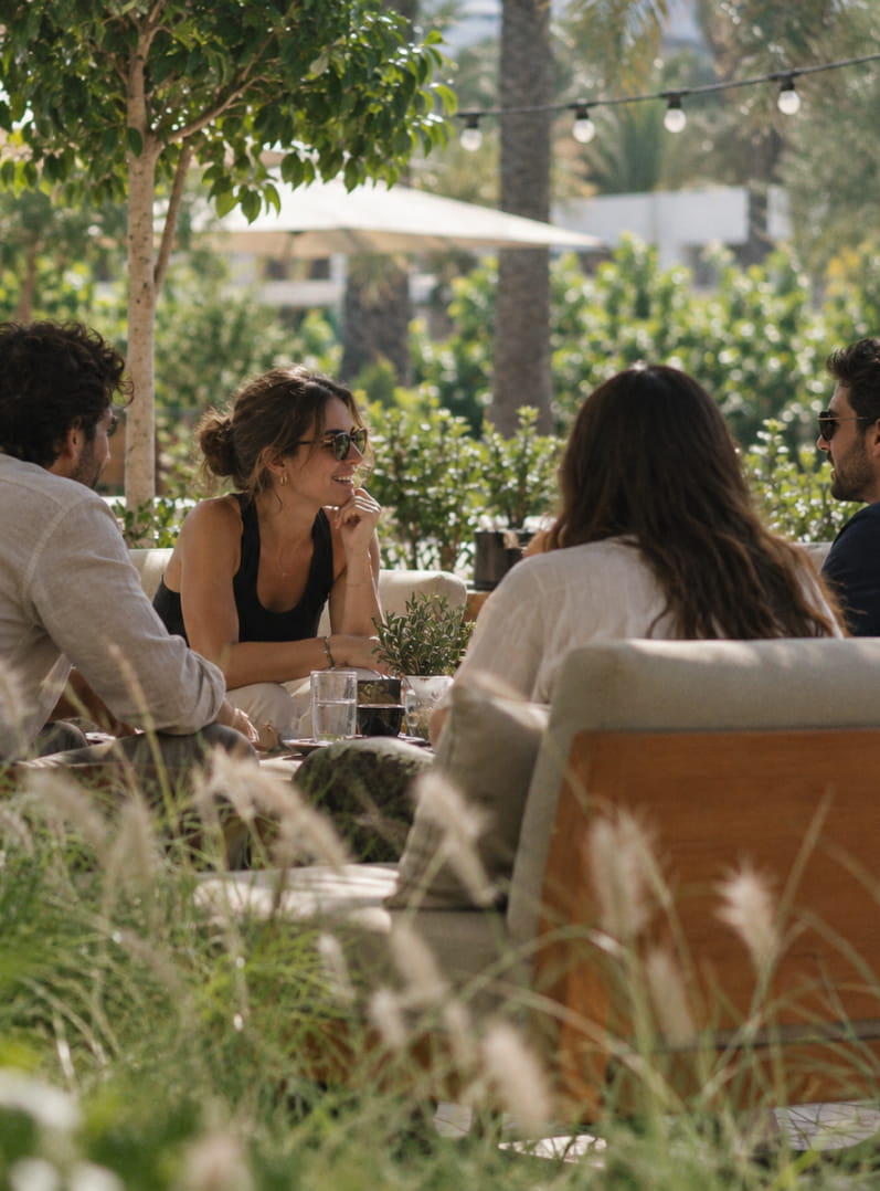 Friends sit around a table in a sunny garden, in the middle of a lively conversation.